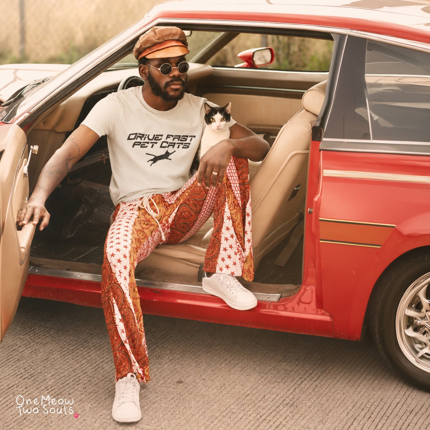 A man sitting in the open door of a red vintage car, wearing a white graphic t-shirt, white sneakers, and distinctive rust-orange paisley patterned pants with white star details. He is holding a black and white cat.
Ivory color Comfort Colors 1717 t-shirt with a "Drive Fast Pet Cats" design. Aimed for Cat dads and men who love cats. One Meow Two Souls brand.