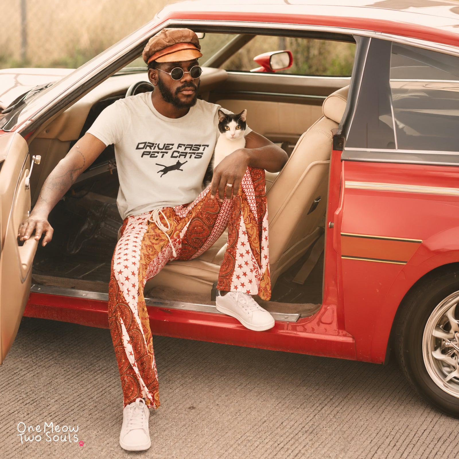 A man sitting in the open door of a red vintage car, wearing a white graphic t-shirt, white sneakers, and distinctive rust-orange paisley patterned pants with white star details. He is holding a black and white cat.
Ivory color Comfort Colors 1717 t-shirt with a "Drive Fast Pet Cats" design. Aimed for Cat dads and men who love cats. One Meow Two Souls brand.