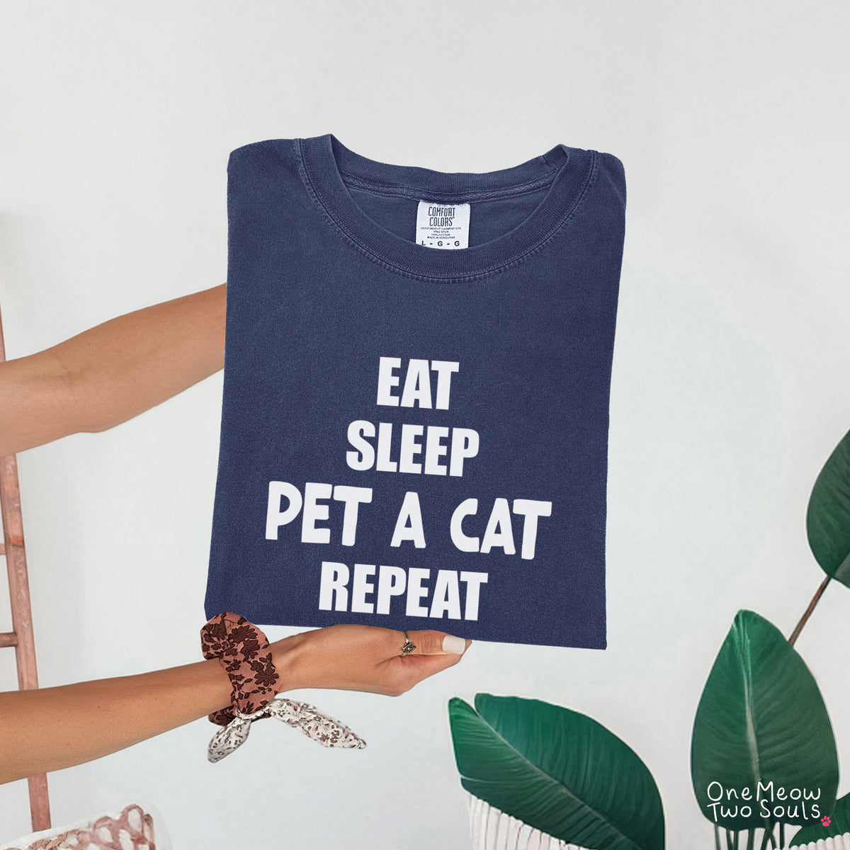 A navy blue folded t-shirt with white text reading 'EAT SLEEP PET A CAT REPEAT' is held by a person's hands against a white wall with green leaves in the foreground.
Navy colored Comfort Colors 1717 t-cotton shirt with a "Eat Sleep Pet a Cat Repeat" design. One Meow Two Souls brand.