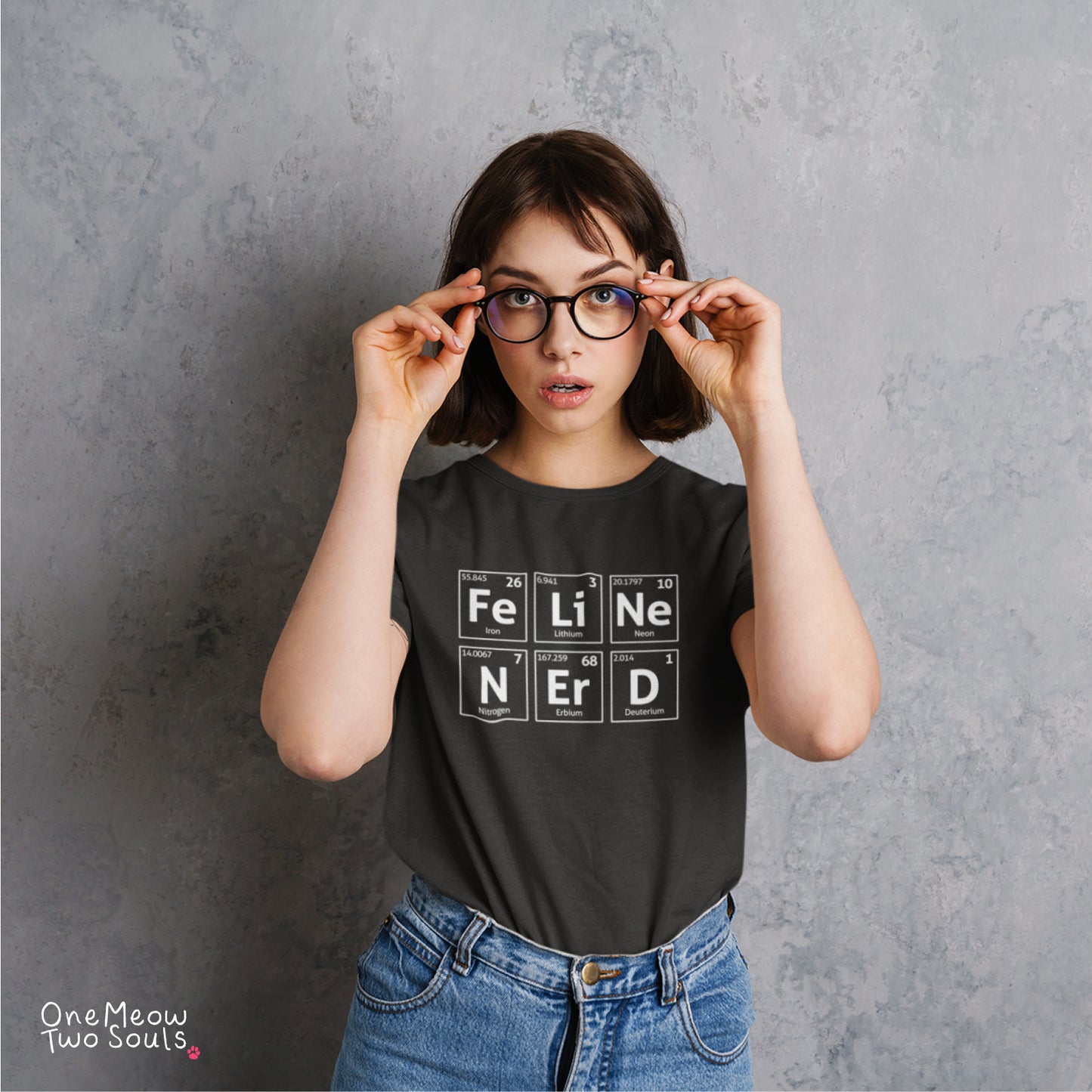 A woman with short brown hair and glasses adjusting her frame, wearing a black graphic t-shirt with periodic table-style blocks spelling 'FELINE NERS' and blue jeans, standing against a textured gray wall. The One Meow Two Souls logo is visible in the bottom-left corner.
