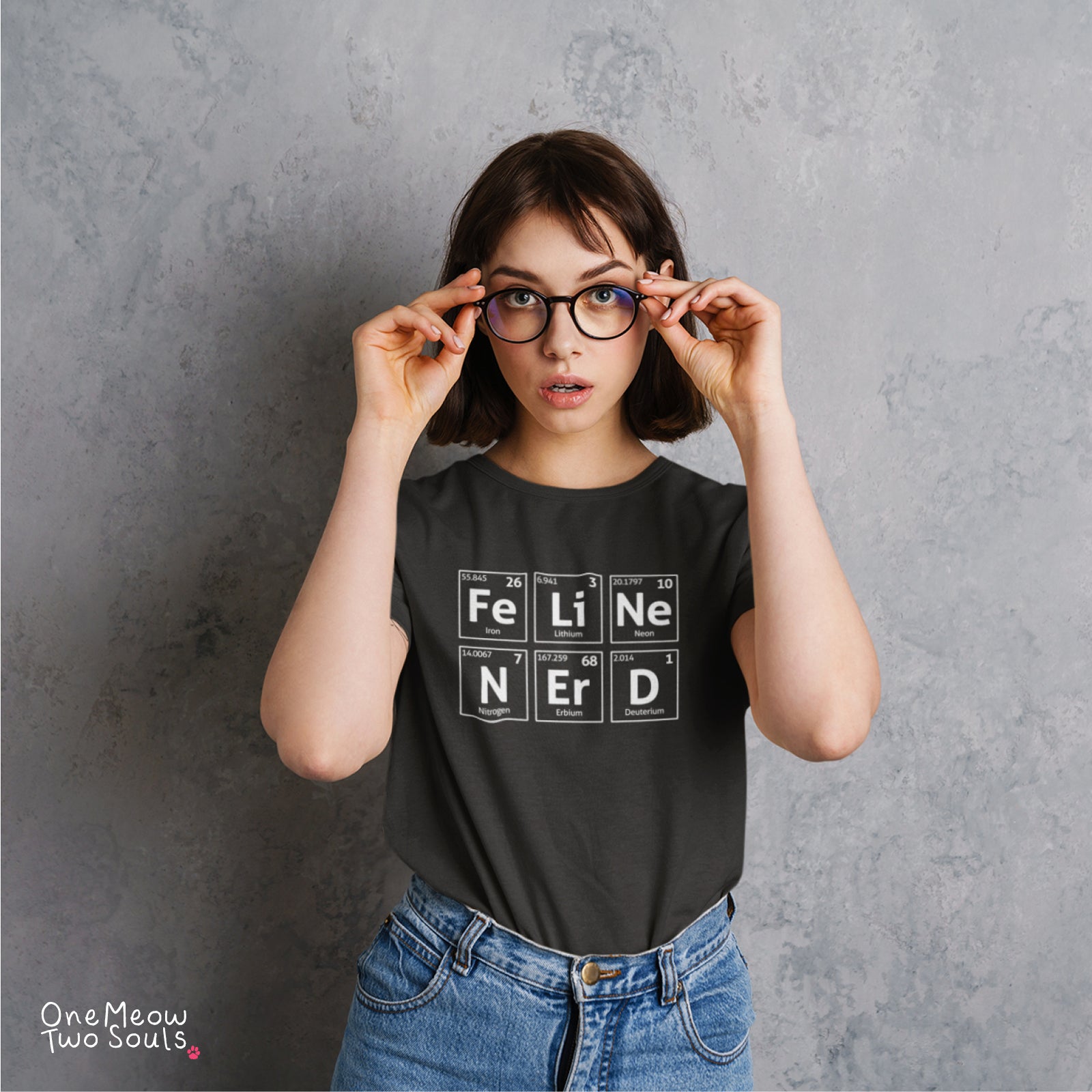 A woman with short brown hair and glasses adjusting her frame, wearing a black graphic t-shirt with periodic table-style blocks spelling 'FELINE NERS' and blue jeans, standing against a textured gray wall. The One Meow Two Souls logo is visible in the bottom-left corner.