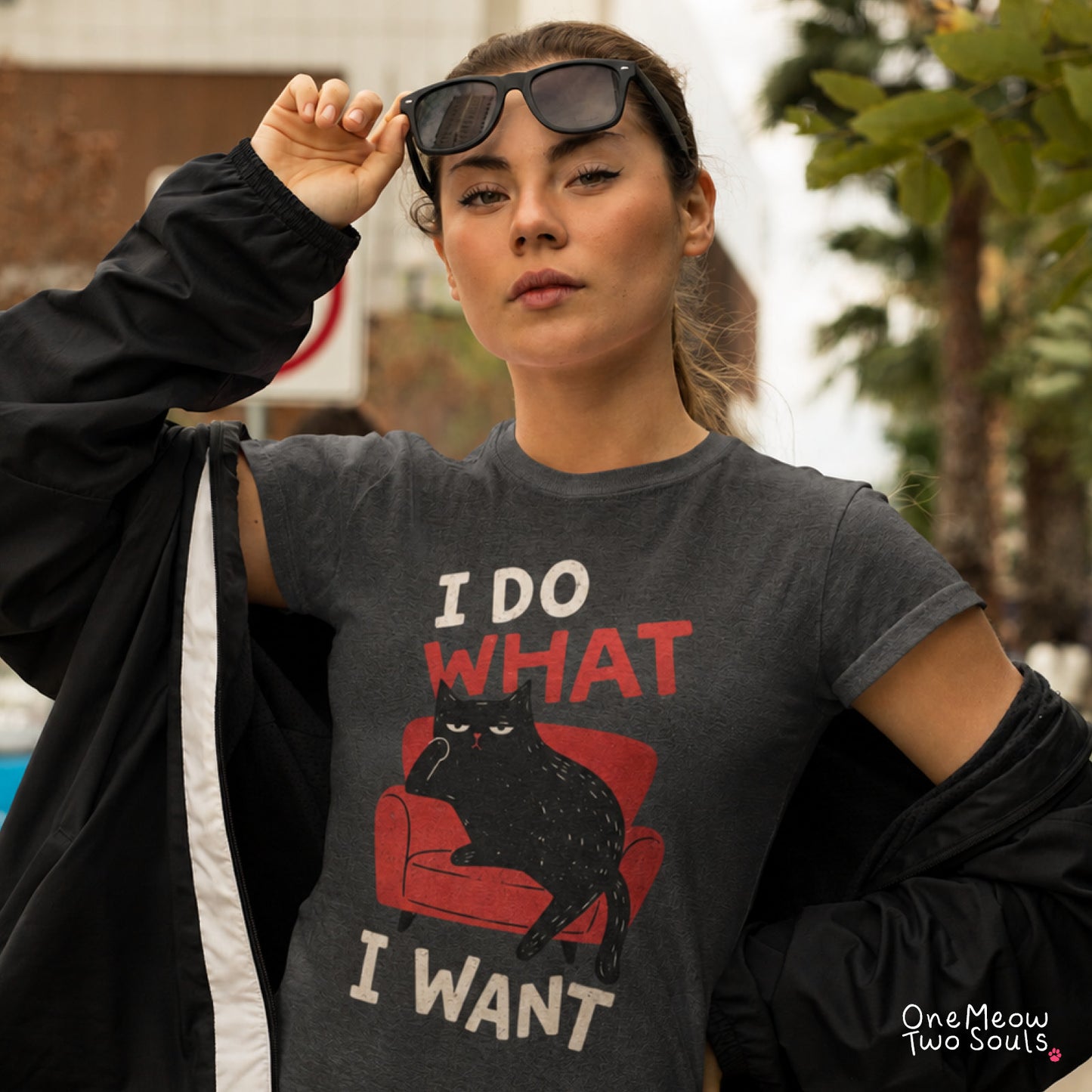 A woman wearing a dark gray graphic t-shirt with a black cat on a red chair and the text 'I DO WHAT I WANT'. She is wearing a black jacket draped off her shoulders and sunglasses on her head, with a blurred outdoor background. One Meow Two Souls Brand.