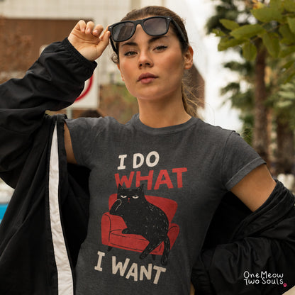 A woman wearing a dark gray graphic t-shirt with a black cat on a red chair and the text 'I DO WHAT I WANT'. She is wearing a black jacket draped off her shoulders and sunglasses on her head, with a blurred outdoor background. One Meow Two Souls Brand.