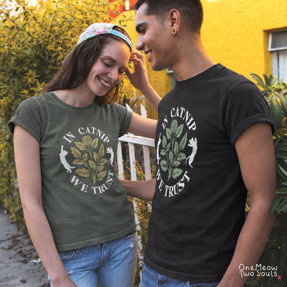 A man and a woman wearing matching graphic t-shirts with the text 'IN CATNIP WE TRUST' and a catnip plant illustration. The woman wears an olive green shirt and a floral cap, while the man wears a black shirt. One Meow Two Souls brand.
