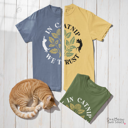 Three graphic t-shirts laid flat on a white wood plank background, with an orange tabby cat curled up near them. The shirts show the phrase “IN CATNIP WE TRUST” with leaf and cat illustrations: one blue shirt, one yellow shirt, and one green shirt.
One Meow Two Souls brand.