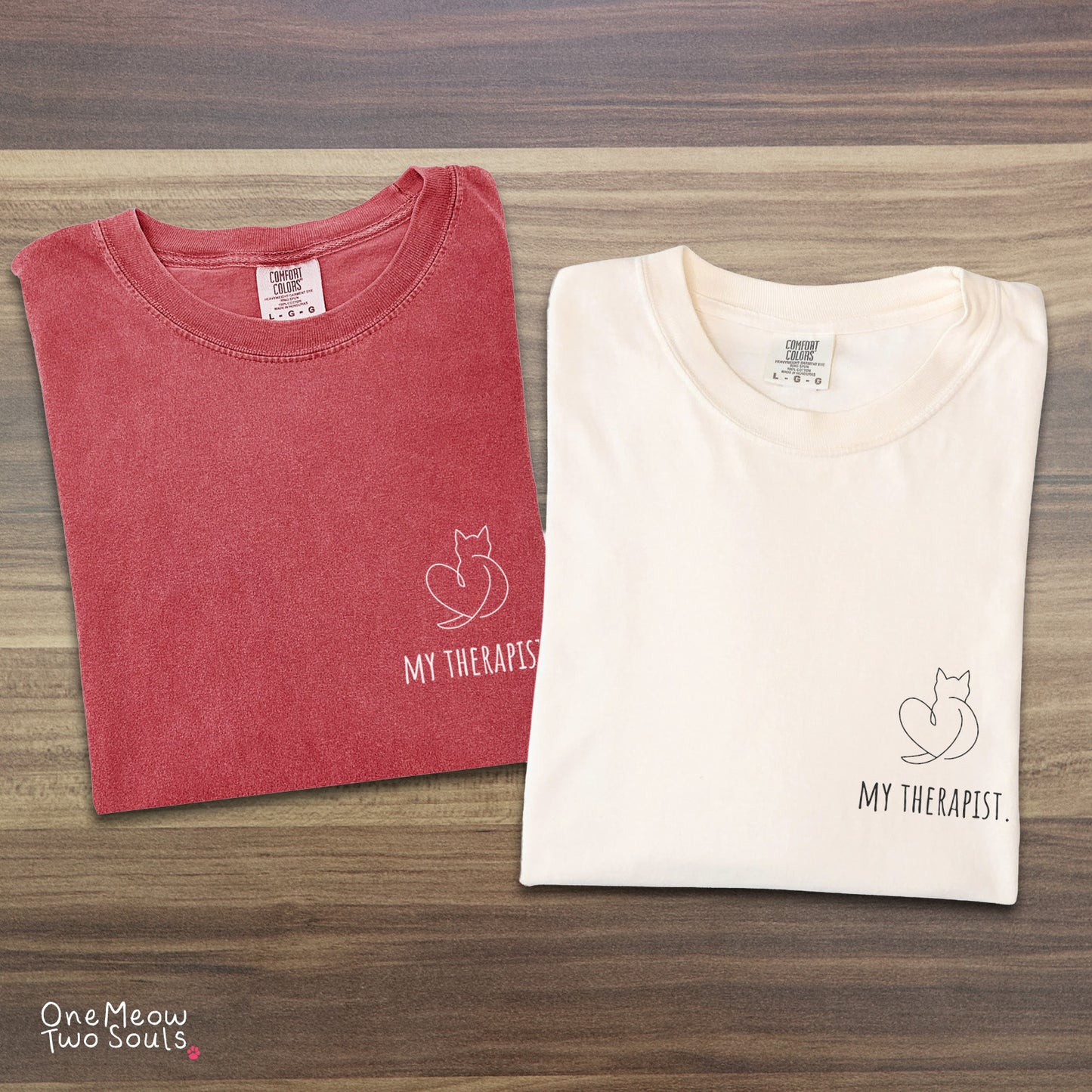 Two folded short-sleeve crewneck t-shirts on a wood-grain surface. The left shirt is a dusty rose red with a black cat outline and the text 'MY THERAPIST.
Ivory and watermelon colored Comfort Colors 1717 t-shirt with a minimal "My Therapist" cat design. One Meow Two Souls brand.