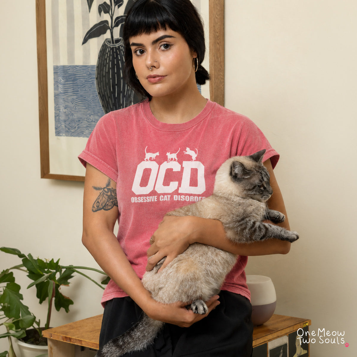 A woman with short dark hair holds a gray and cream cat in her lap while wearing a pink t-shirt with a white graphic that reads 'OCD OBSESSIVE CAT DISORDER'. The background shows a warm indoor setting with a framed print, a wooden bench, and a potted plant.
Watermelon colored Comfort Colors 1717 t-shirt with a "OCD - Obsessive Cat Disorder" design. One Meow Two Souls brand.