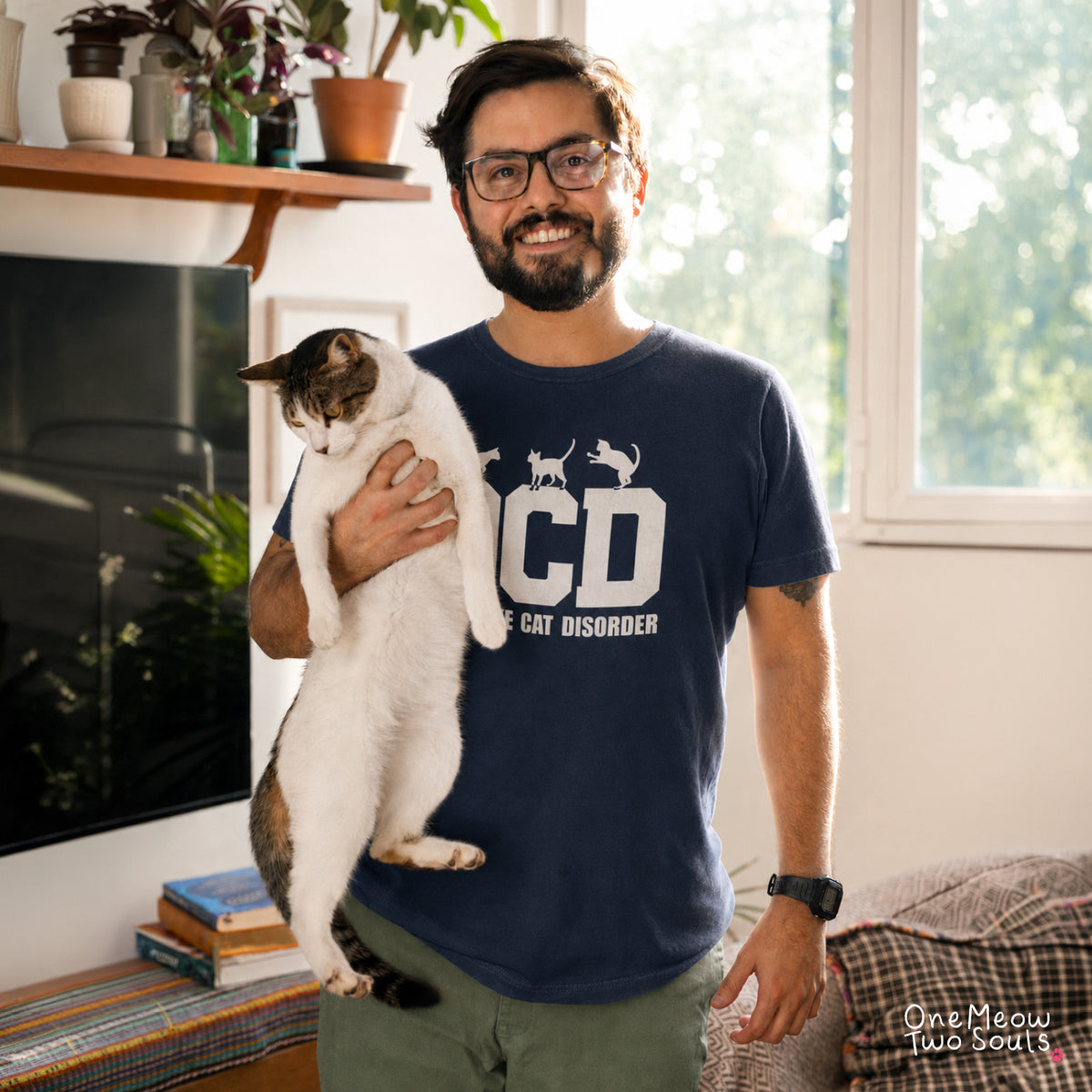 A man wearing glasses and a navy blue graphic t-shirt with a cat, standing in a bright living room. The t-shirt features a white graphic of a cat and the text 'OCD' and 'THE CAT DISORDER'.
Navy colored Comfort Colors 1717 t-shirt with a "OCD - Obsessive Cat Disorder" design. One Meow Two Souls brand.