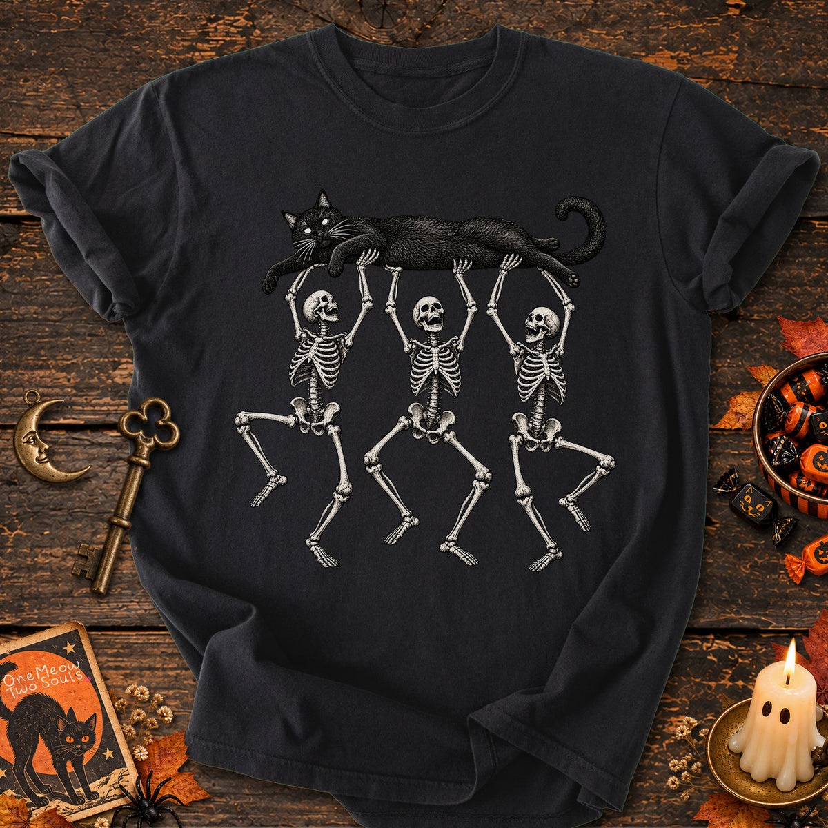 Black colored Comfort Colors 1717 cotton T-shirt, featuring a Black cat carried by three skeletons funny cat Halloween design illustration. One Meow Two Souls brand.