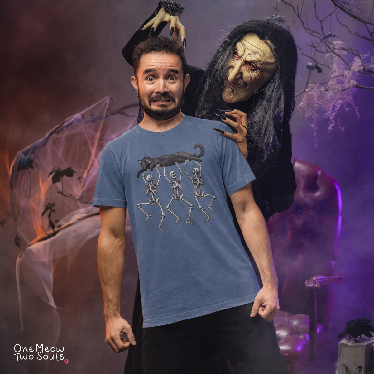 Blue jean colored Comfort Colors 1717 cotton T-shirt, featuring a Black cat carried by three skeletons funny cat Halloween design illustration. One Meow Two Souls brand.