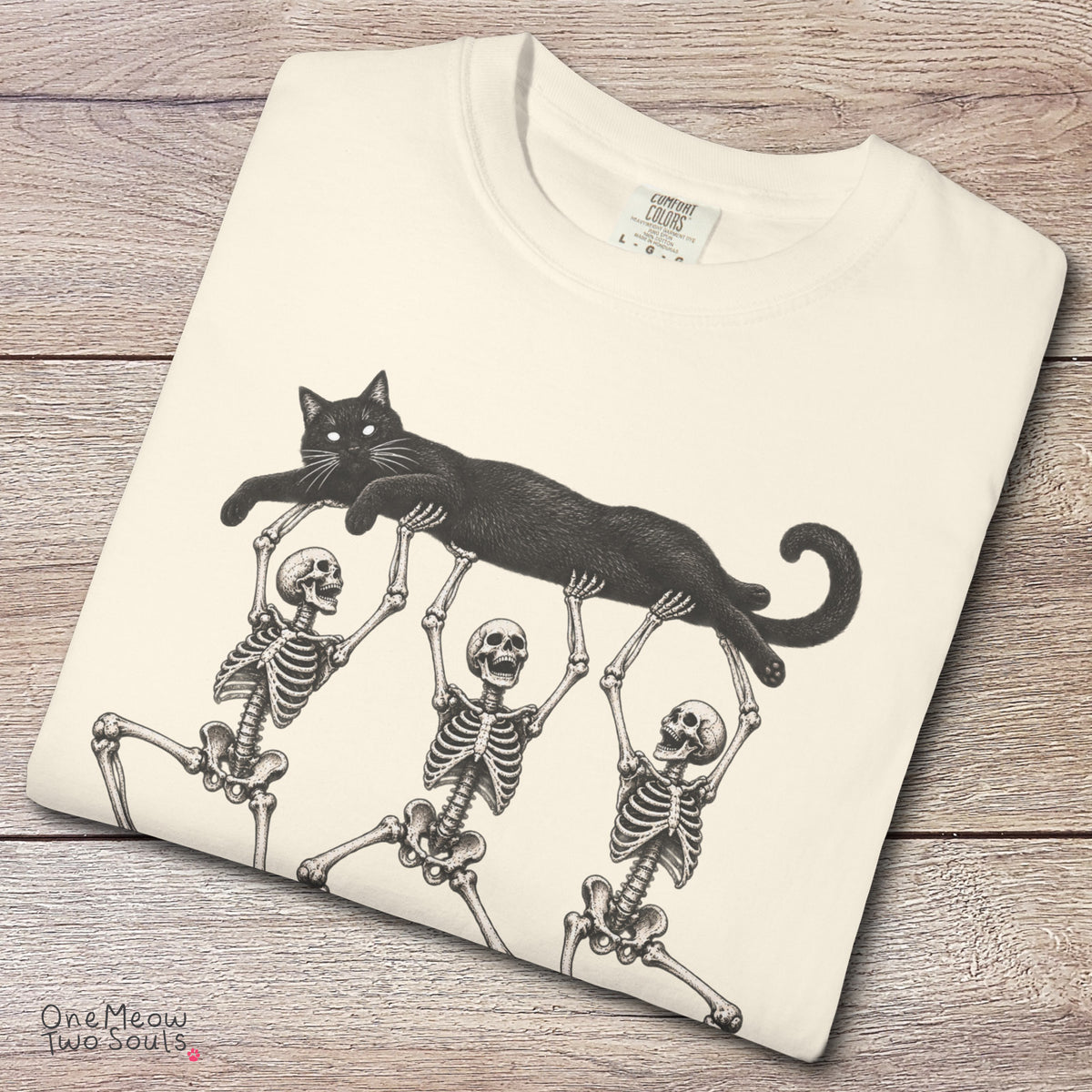 Ivory colored Comfort Colors 1717 cotton T-shirt, featuring a Black cat carried by three skeletons funny cat Halloween design illustration. One Meow Two Souls brand.