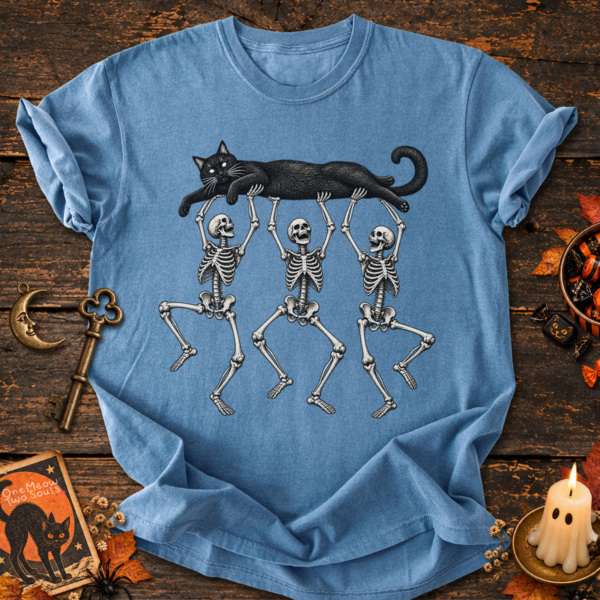 Blue jean colored Comfort Colors 1717 cotton T-shirt, featuring a Black cat carried by three skeletons funny cat Halloween design illustration. One Meow Two Souls brand.