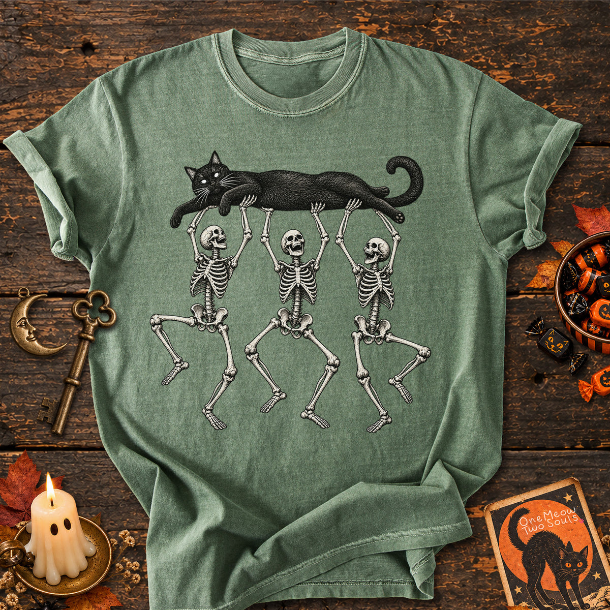 Moss colored Comfort Colors 1717 cotton T-shirt, featuring a Black cat carried by three skeletons funny cat Halloween design illustration. One Meow Two Souls brand.