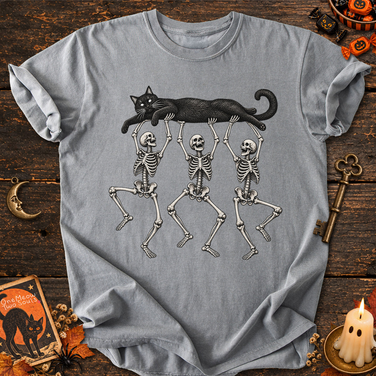 Gray colored Comfort Colors 1717 cotton T-shirt, featuring a Black cat carried by three skeletons funny cat Halloween design illustration. One Meow Two Souls brand.