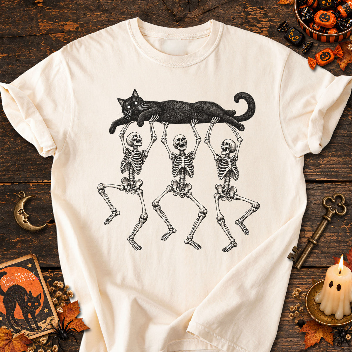 Ivory colored Comfort Colors 1717 cotton T-shirt, featuring a Black cat carried by three skeletons funny cat Halloween design illustration. One Meow Two Souls brand.