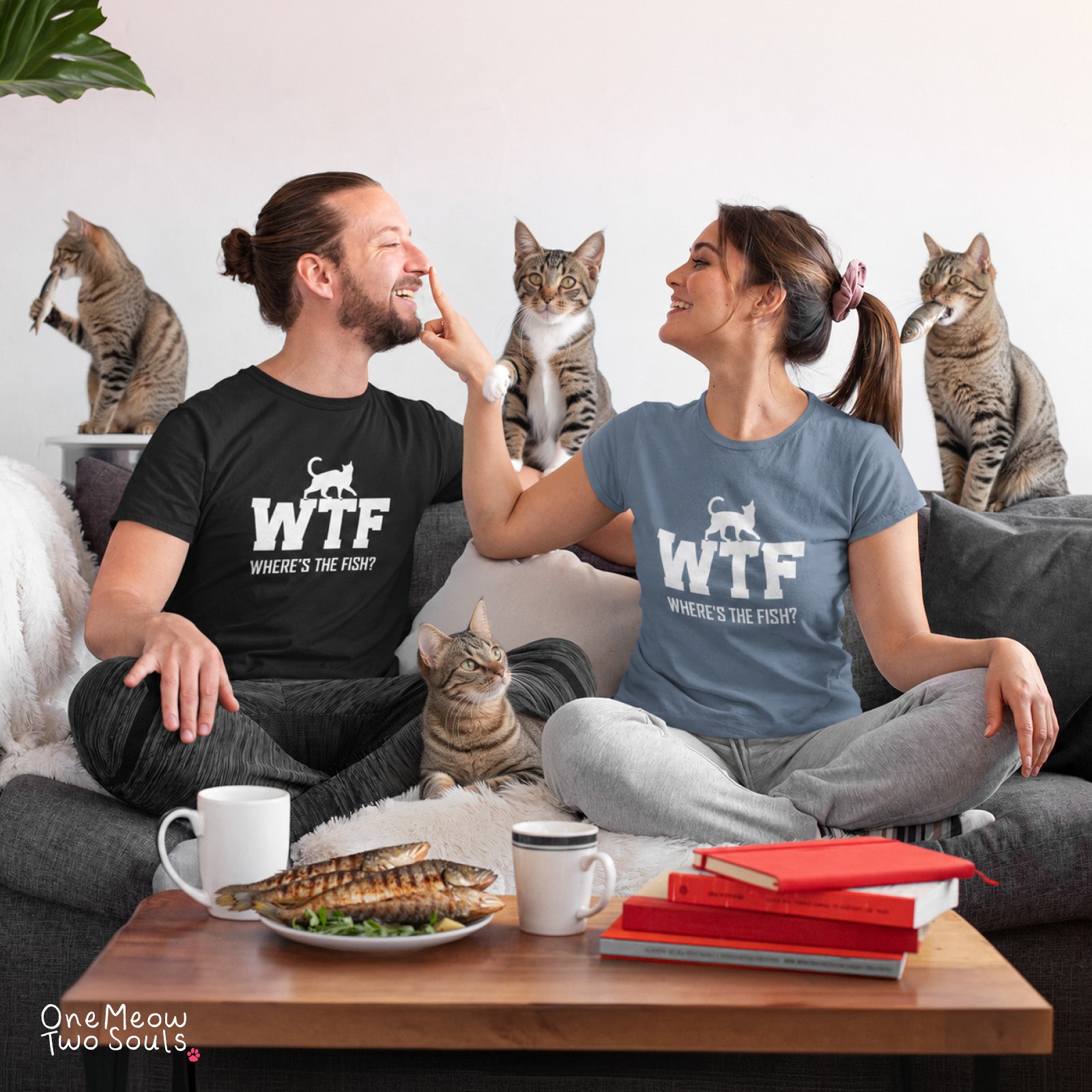 A man and a woman sitting on a couch wearing matching graphic t-shirts that read 'WTF WHERE'S THE FISH?' with a cat silhouette. Several tabby cats are sitting on the couch and table.
Comfort Colors 1717 t-shirts presenting a "WTF Where's The Fish?" funny cat design. One Meow Twou Sould brand.