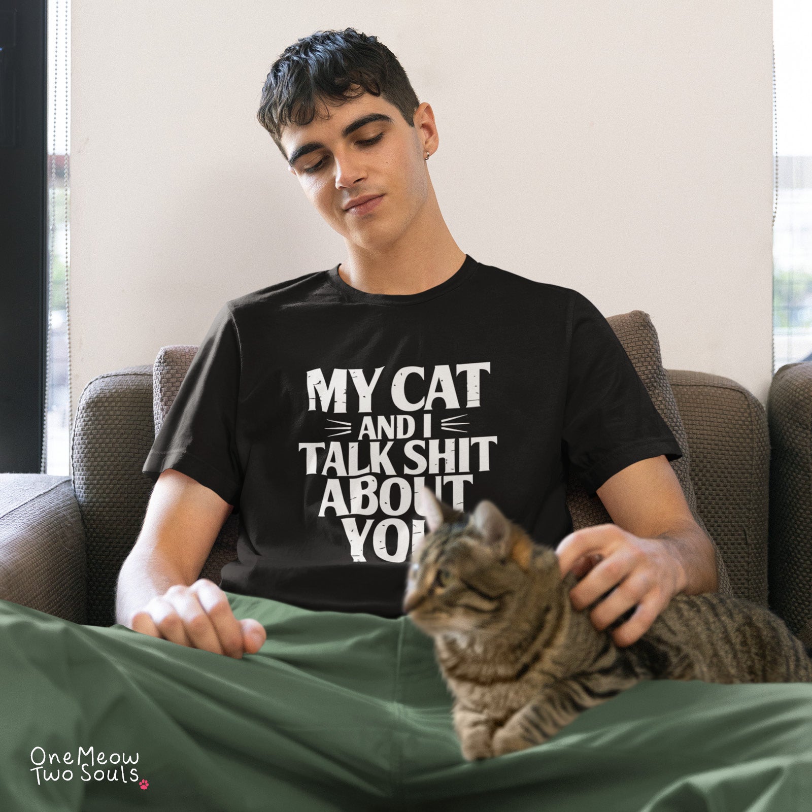 A man sitting on a couch wearing a black graphic t-shirt that reads 'MY CAT AND I TALK SHIT ABOUT YOU' in white text, with a brown tabby cat resting on his lap. The photo is taken in a home setting with a neutral background. One Meow Two Souls brand.