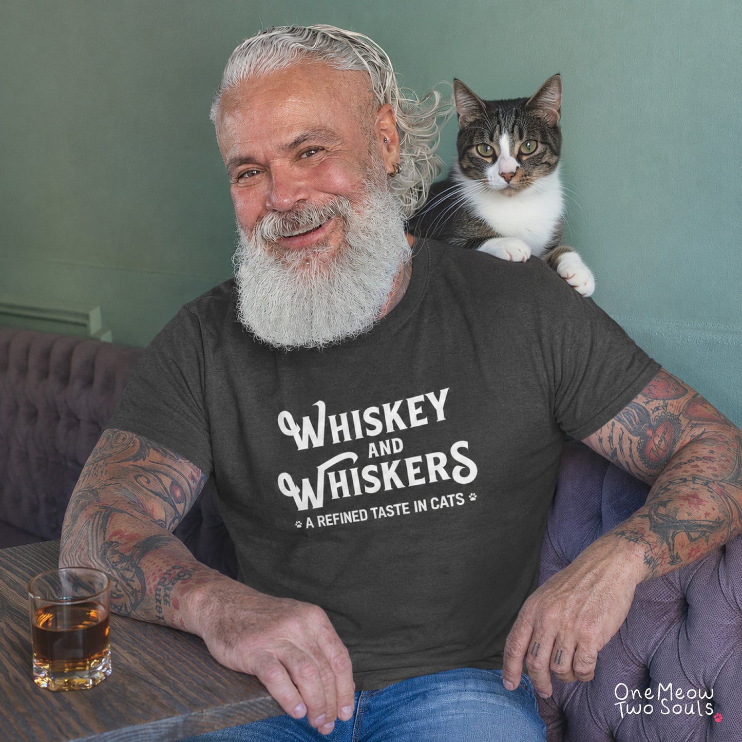 A smiling man with a long white beard and tattoos sits in a purple tufted armchair, wearing a dark charcoal t-shirt with the white text 'Whiskey and Whiskers - A Refined Taste in Cats'. A tabby and white cat rests on his shoulder.
Pepper colored Comfort Colors 1717 cotton T-shirt, with a "Whiskey and Whiskers" design. One Meow Two Souls brand.