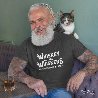A smiling man with a long white beard and tattoos sits in a purple tufted armchair, wearing a dark charcoal t-shirt with the white text 'Whiskey and Whiskers - A Refined Taste in Cats'. A tabby and white cat rests on his shoulder.
Pepper colored Comfort Colors 1717 cotton T-shirt, with a "Whiskey and Whiskers" design. One Meow Two Souls brand.