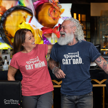 A smiling woman in a coral pink t-shirt and a bearded man in a navy blue t-shirt, both featuring the white printed text 'World's Dopest CAT Mom / CAT Dad'. They are standing in a bar environment with a colorful mural in the background. One Meow Two Souls brand.