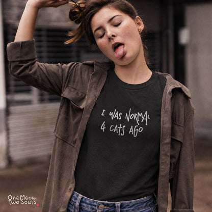 A woman wearing a black graphic t-shirt with the text 'I WAS NORMAL 4 CATS AGO' in white handwritten font, layered under an open brown suede jacket. She is posing in an urban setting with a blurred background. One Meow Two Souls.