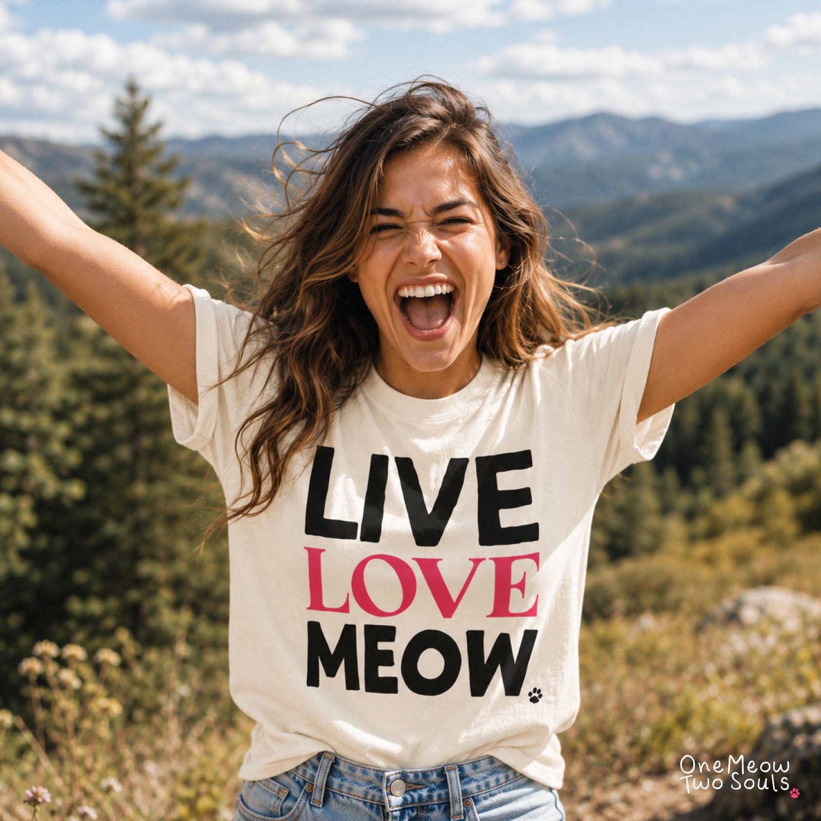 A woman outdoors with her arms raised, wearing a cream graphic T-shirt that reads 'LIVE LOVE MEOW' in black and pink lettering, paired with blue jeans. Mountain and forest scenery is visible in the background. One Meow Two Souls brand.