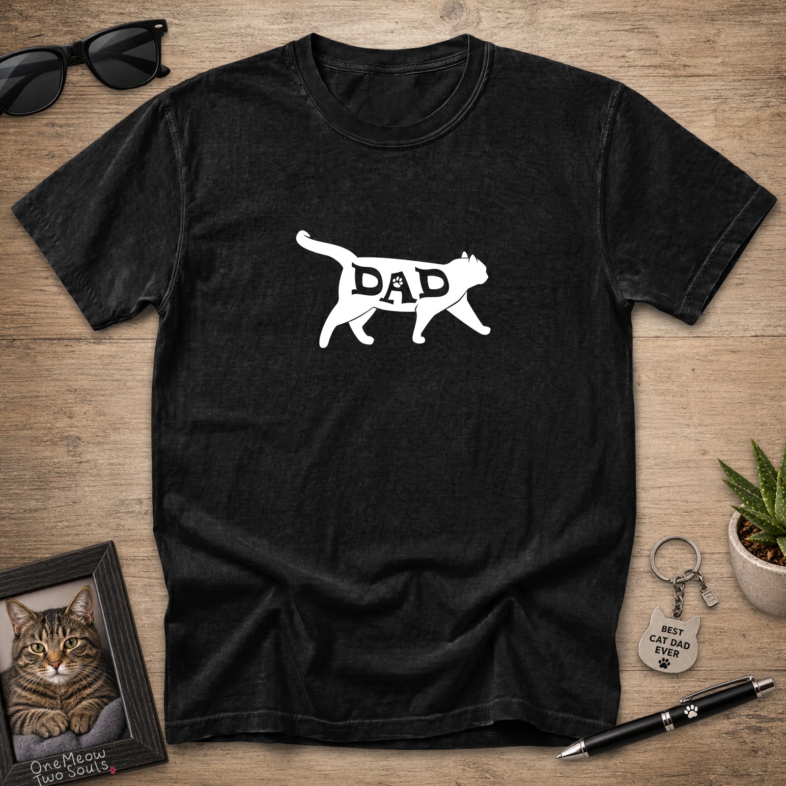 A black short-sleeve t-shirt with a white graphic of a cat silhouette containing the word 'DAD', displayed in a flat-lay arrangement on a wooden surface with sunglasses, a framed cat photo, a small plant, and a pen.