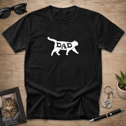A black short-sleeve t-shirt with a white graphic of a cat silhouette containing the word 'DAD', displayed in a flat-lay arrangement on a wooden surface with sunglasses, a framed cat photo, a small plant, and a pen.
