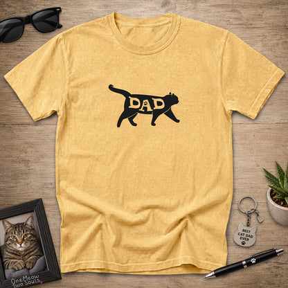 A heathered mustard yellow t-shirt laid flat on a wooden surface, featuring a black silhouette of a walking cat with the word 'DAD' filled in with gold lettering. The shirt is styled with sunglasses, a framed picture of a cat, a small plant, and a pen.