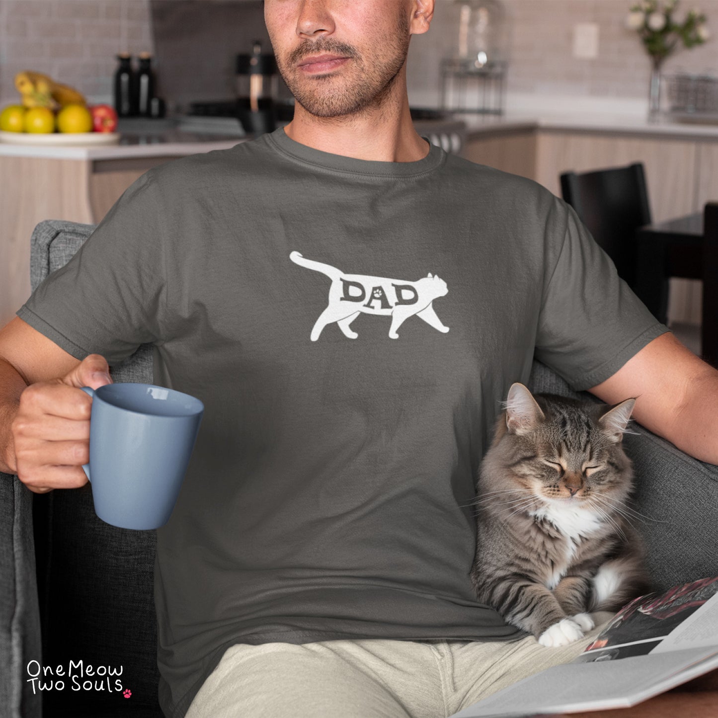 A man wearing a dark gray t-shirt with a white graphic of a cat silhouette containing the word 'DAD' sits in a living room setting, holding a blue mug with a tabby cat resting on his lap.