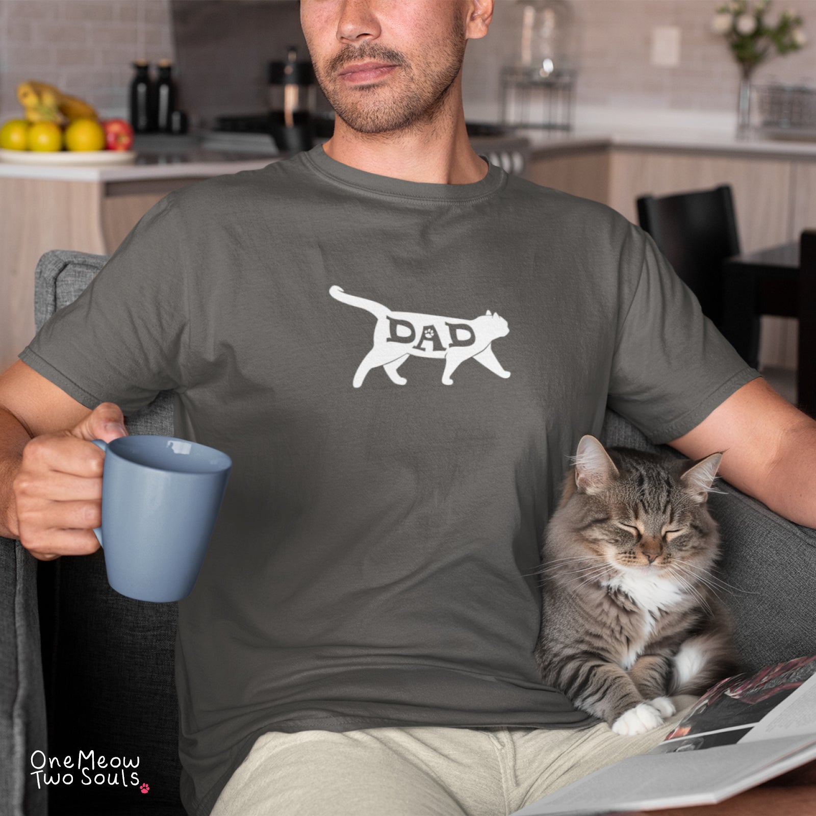 A man wearing a dark gray t-shirt with a white graphic of a cat silhouette containing the word 'DAD' sits in a living room setting, holding a blue mug with a tabby cat resting on his lap.