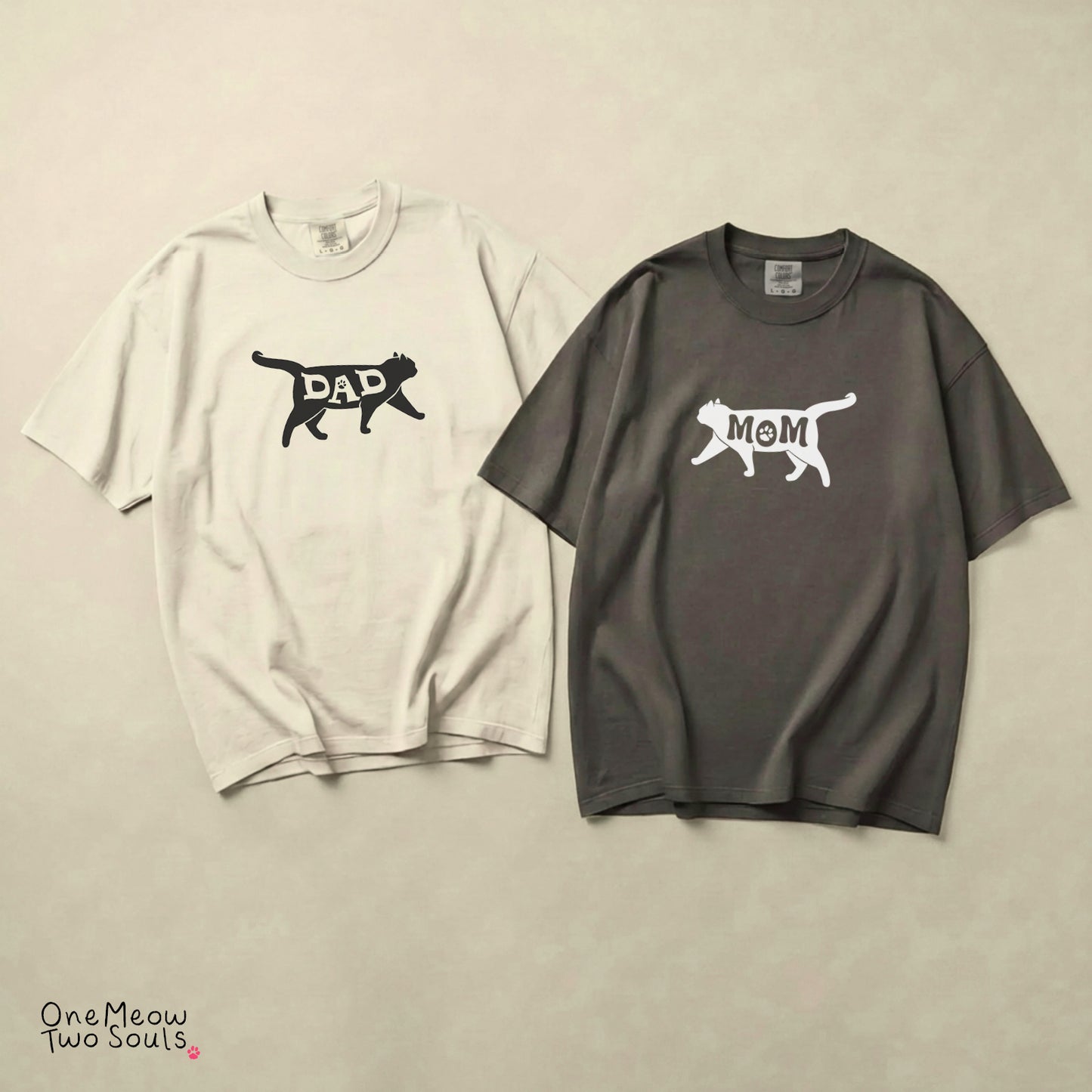 Two oversized short-sleeve T-shirts laid flat on a light beige textured background. The left shirt is off-white with a black cat silhouette graphic containing the word 'DAD'. The right side says 'MOM', both with a silhouette of a cat.