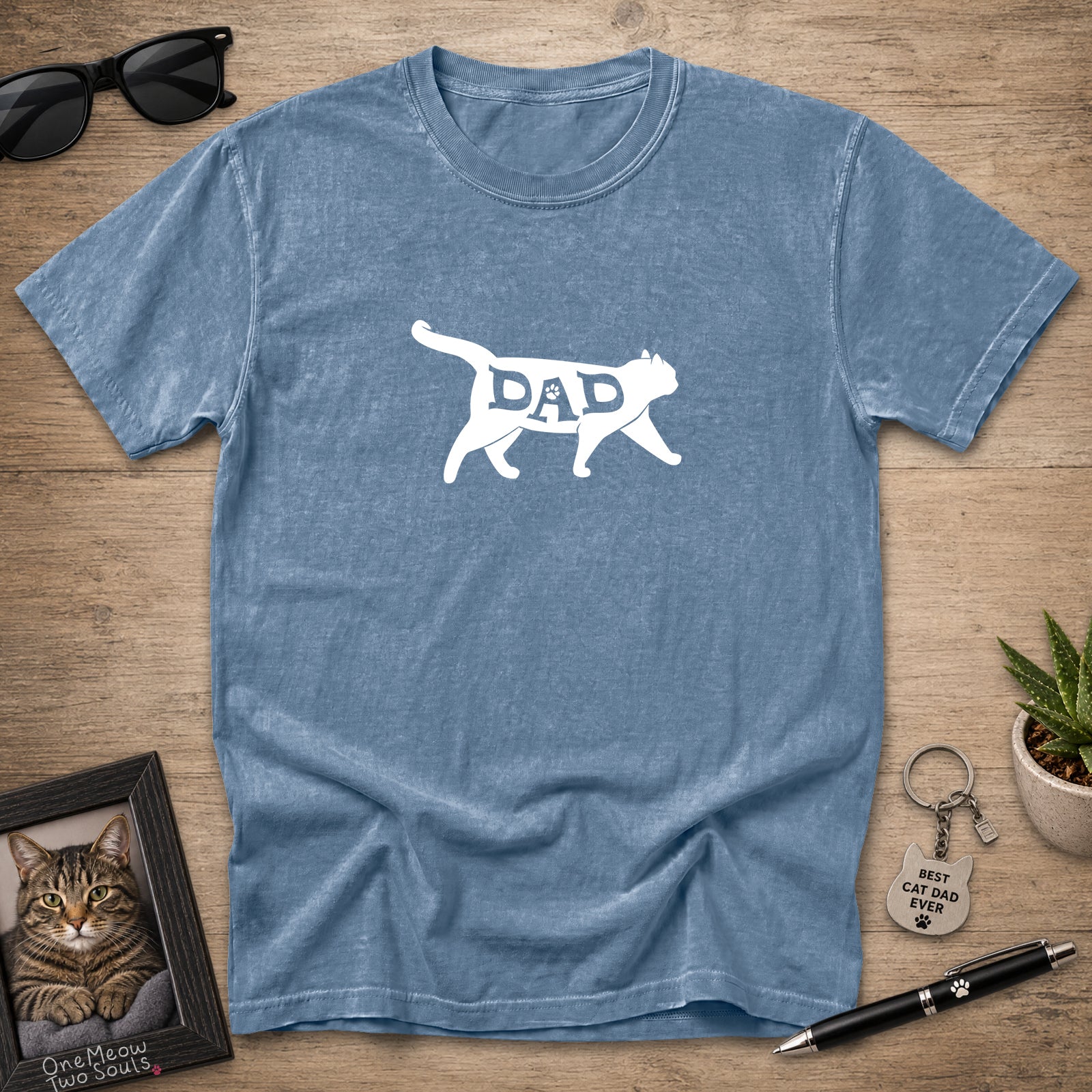 A heathered blue short-sleeve t-shirt laid flat on a wooden surface, featuring a white silhouette of a walking cat with the word 'DAD' inside. Sunglasses, a framed cat portrait, a small plant, and a keychain are arranged around it as styling props.