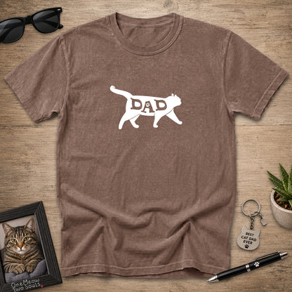 A brown short-sleeve crewneck t-shirt laid flat on a wooden surface, featuring a white graphic of a cat silhouette with the word 'DAD' inside. The shirt is styled with sunglasses, a framed cat portrait, a small succulent, and a pen.