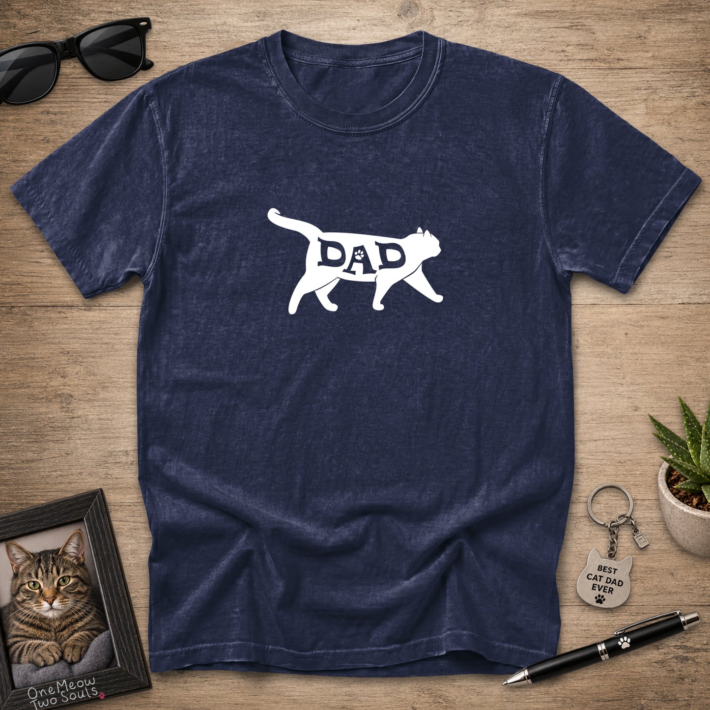 A navy blue short-sleeve t-shirt with a white graphic of a walking cat containing the word 'DAD' inside its body, displayed on a wooden tabletop flat-lay with sunglasses, a framed cat portrait, a small plant, and a pen.