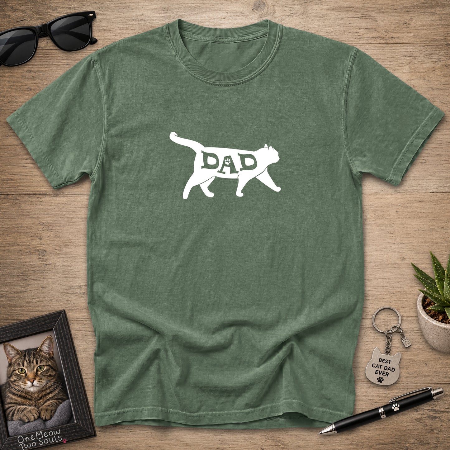 A heathered green crewneck t-shirt with a white graphic of a cat silhouette containing the word 'DAD', displayed in a flat-lay arrangement on a wooden surface with sunglasses, a framed cat portrait, a keychain, and a pen.