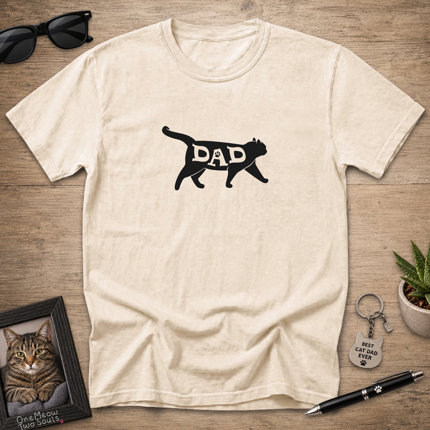 A cream-colored short-sleeve t-shirt with a black graphic of a cat silhouette containing the word 'DAD' in white, laid flat on a wooden surface with sunglasses, a framed cat photo, a keychain, and a pen as props.