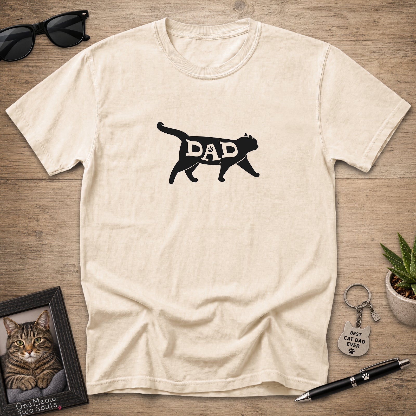 A cream-colored short-sleeve t-shirt with a black graphic of a cat silhouette containing the word 'DAD' in white, laid flat on a wooden surface with sunglasses, a framed cat photo, a keychain, and a pen as props.