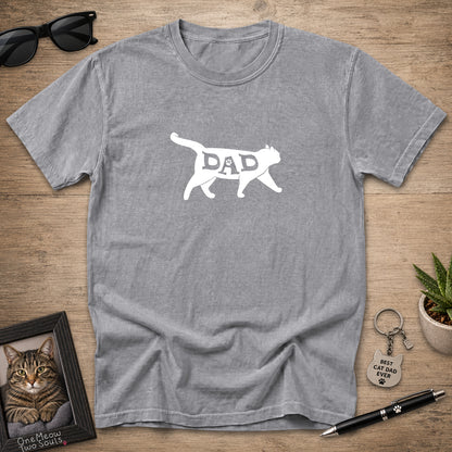 A heather gray short-sleeve t-shirt laid flat on a wooden surface, featuring a white silhouette of a cat with the word 'DAD' inside it. The shirt is styled with sunglasses, a framed cat portrait, a small plant, a keychain, and a pen.
