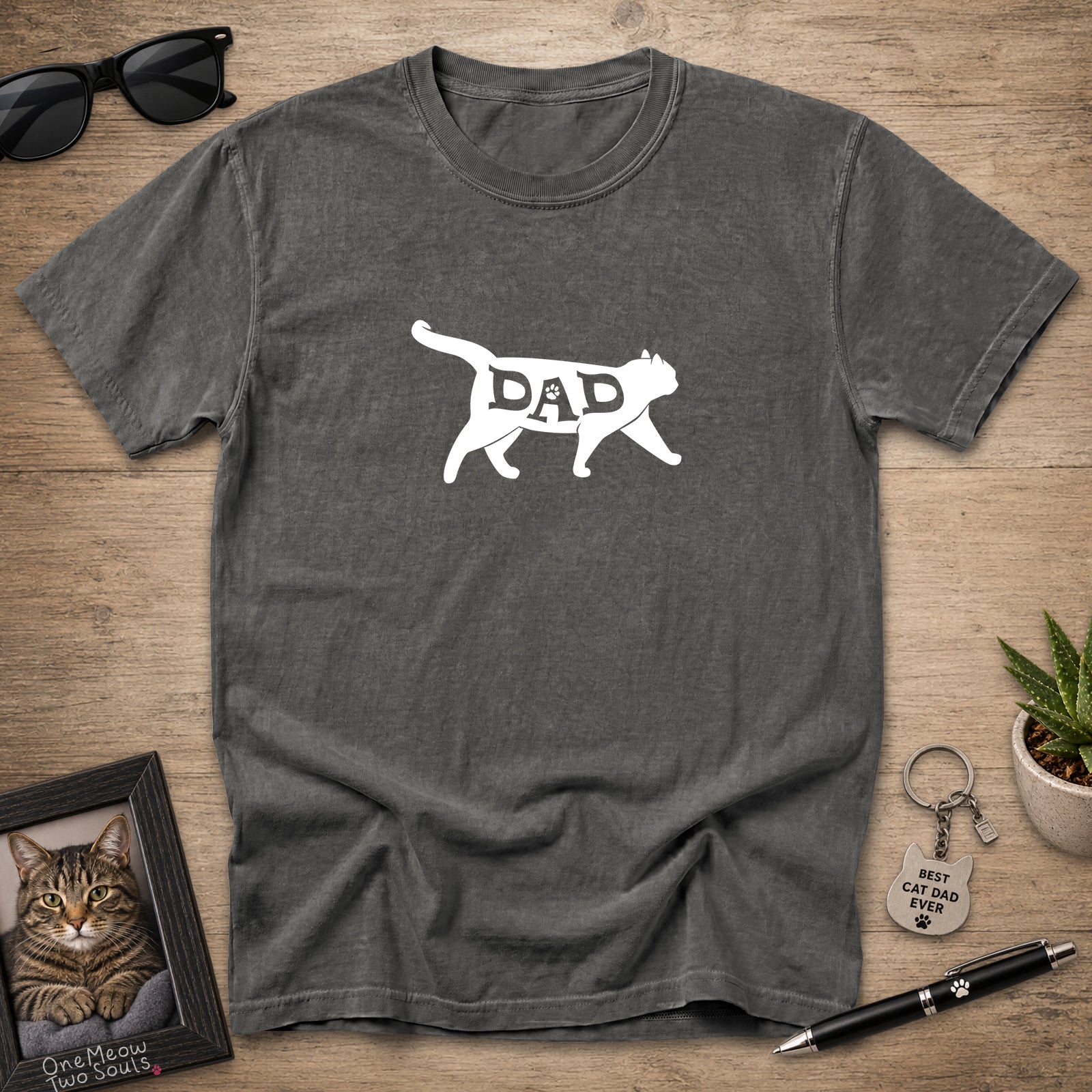 A dark gray heather t-shirt with a white graphic of a cat silhouette containing the word 'DAD' inside, laid flat on a wooden surface with sunglasses, a framed cat portrait, a small plant, and a pen around it.