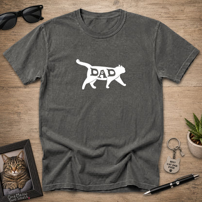 A dark gray heather t-shirt with a white graphic of a cat silhouette containing the word 'DAD' inside, laid flat on a wooden surface with sunglasses, a framed cat portrait, a small plant, and a pen around it.