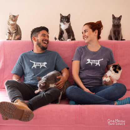 A man and a woman sitting on a pink couch wearing matching gray t-shirts with white dog silhouette graphics labeled 'DAD' and 'MOM'. Cats are present on and around the couch, and the photo has a small 'One Meow Two Souls' mark in the bottom right corner.