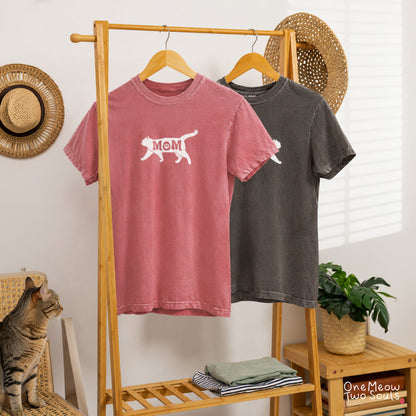 Two short-sleeve graphic T-shirts hanging on a wooden clothing rack in a styled room. The front shirt is Comfort Colors 1717 Watermelon color shirt with a white cat silhouette and the word 'MOM' printed on the chest. One Meow Two Souls brand.